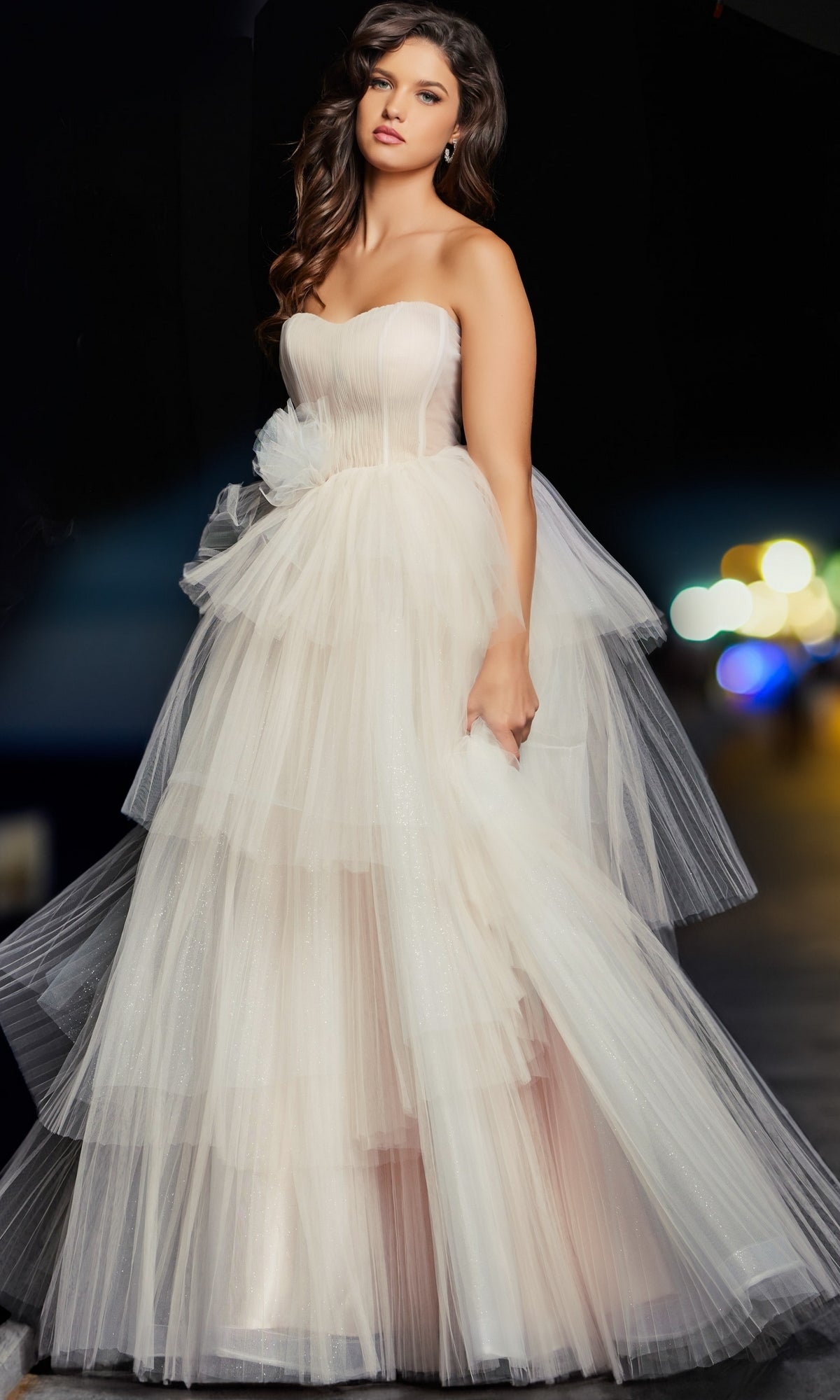 A woman in the Jovani Blush Pink Long Prom Ball Gown 38537, featuring a strapless sweetheart corset bodice and layered tulle skirt, stands against a dark background with blurred lights.